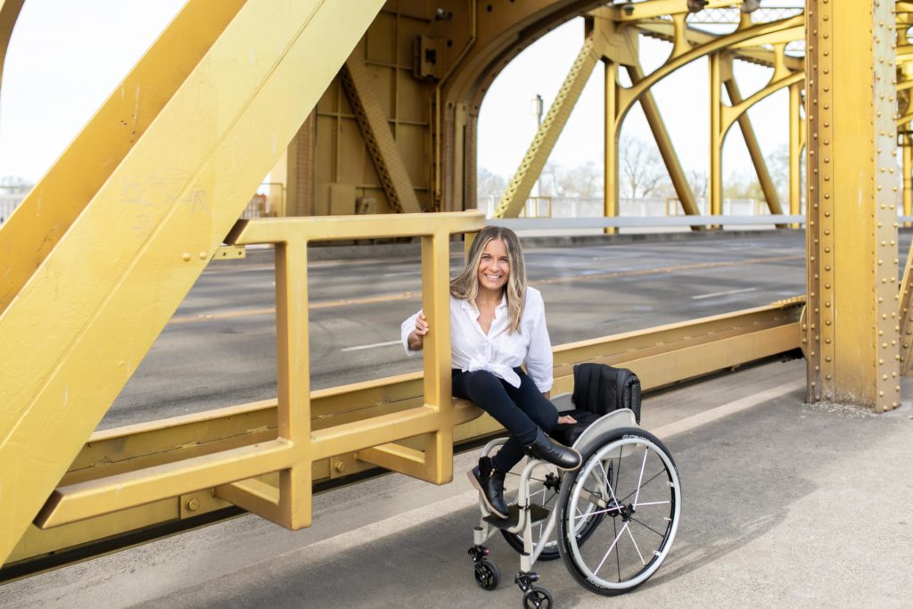 Alycia sits on a yellow bridge with her hand and foot on the wheelchair next to her