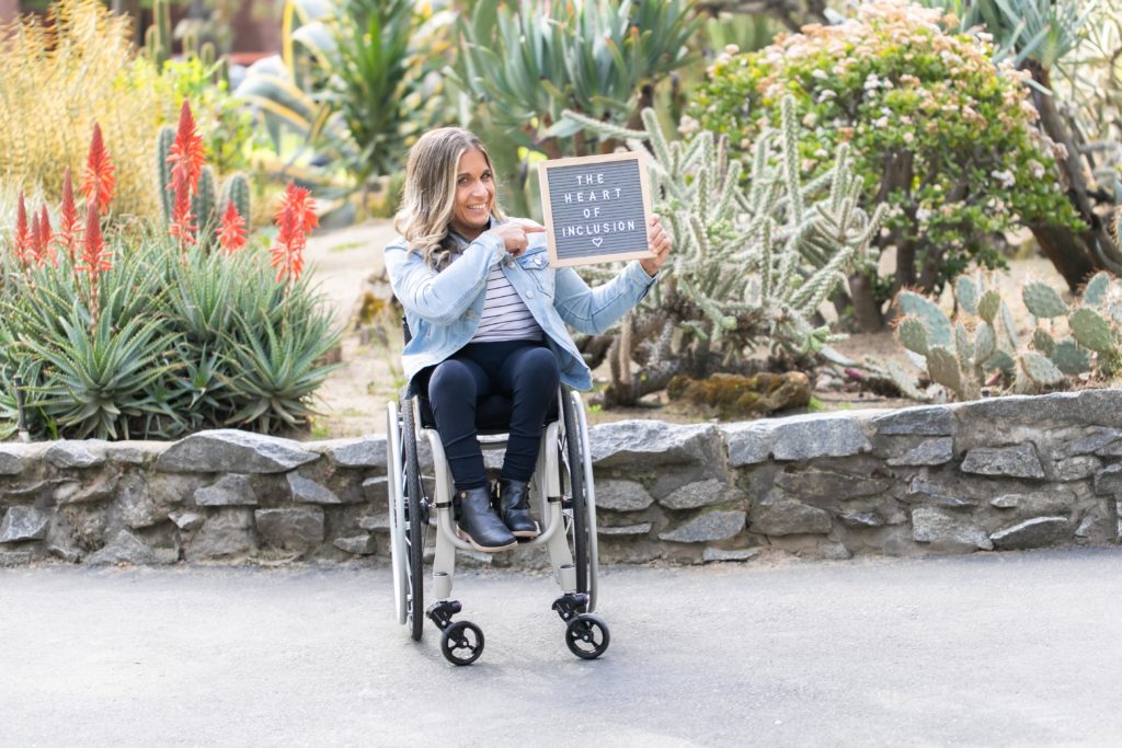 With exotic plants behind her, Alycia in her wheelchair holds a sign "The heart of inclusion"