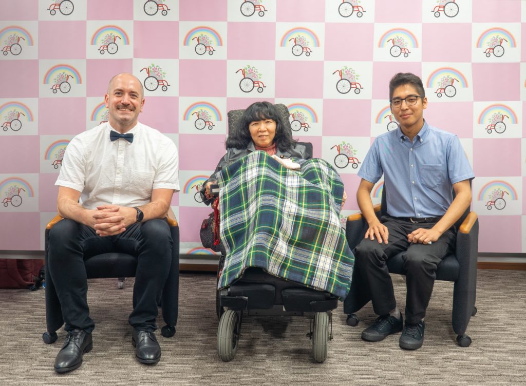 トマスローガンが電動車椅子にのる木村英子氏の左にこちらを向いて座っている。反対の右側には著者のやながわけんじが座っている。