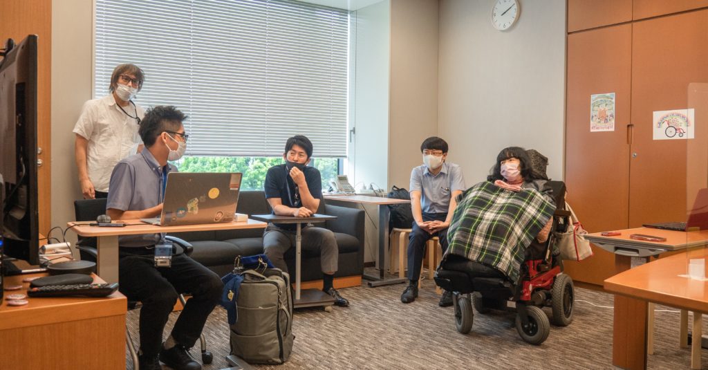 Kenji sits with his laptop facing Eiko Kimura who is in a wheelchair with three others behind them