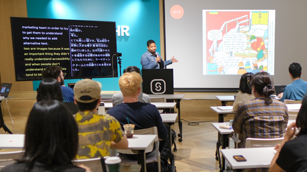 Reginé Gilbert at the podium holding a microphone. To her left is a screen with her presentation. To her right is a large monitor that shows captions in English on one side and Japanese on the other side.