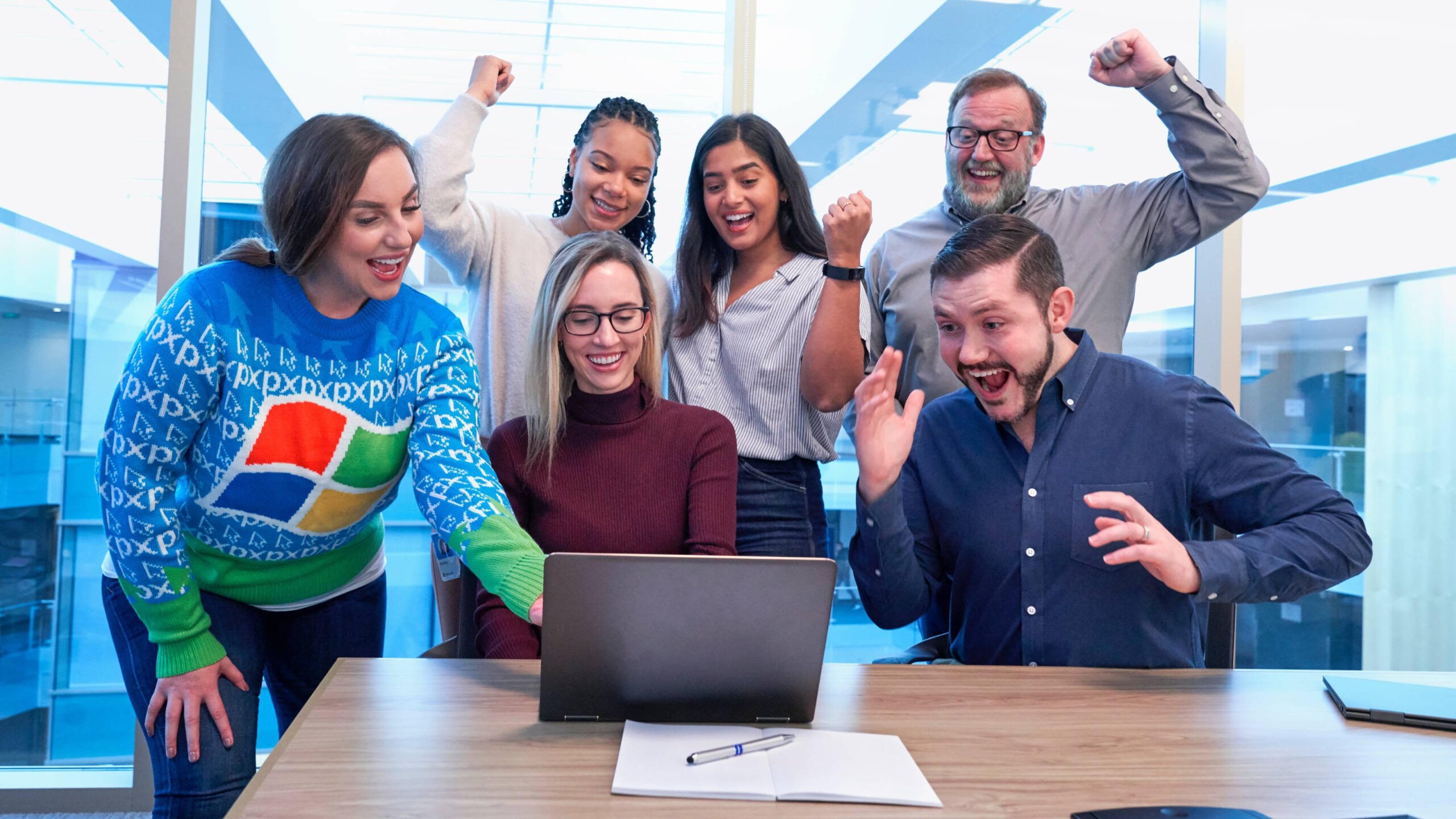 Six people gathered around a laptop, celebrating with joyful expressions. One person wears a colorful sweater with the Windows logo, while others are smiling, cheering, and raising their hands in excitement.