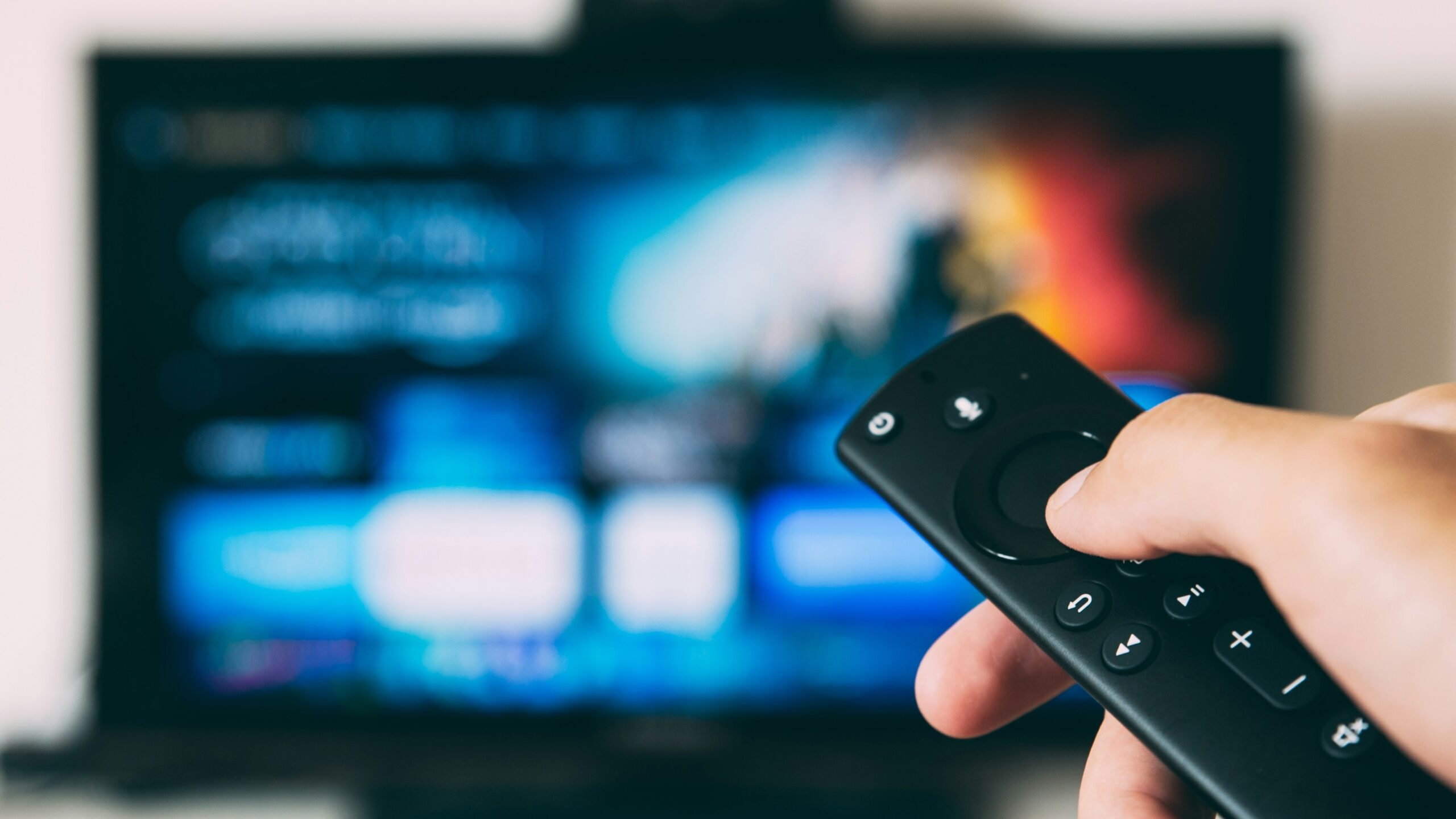 A pale-skinned hand pointing a remote at a blurry TV some distance ahead.