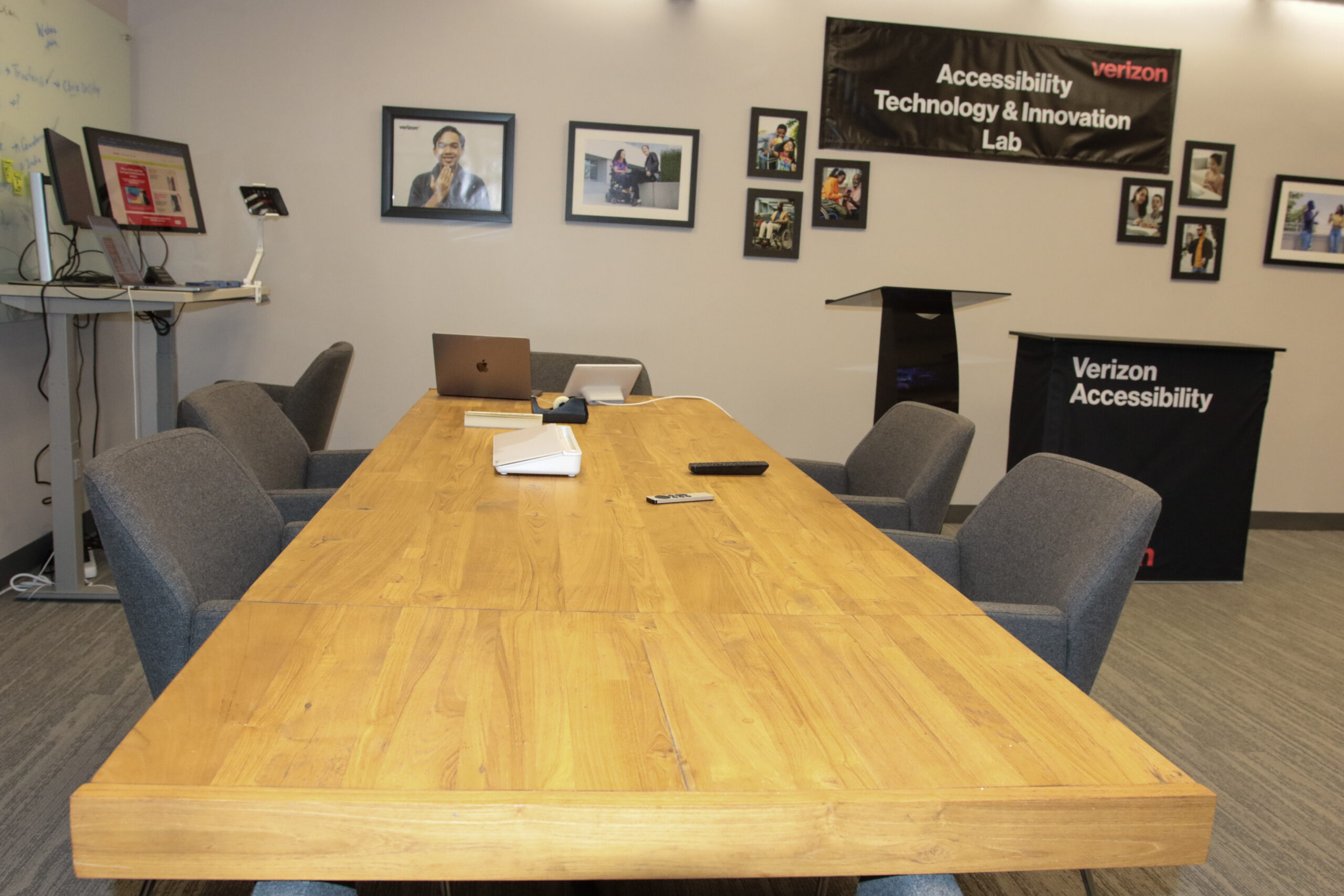 Collaboration space with a long wooden table, five chairs, and a laptop. A computer standing desk is on the left and a podium on the right