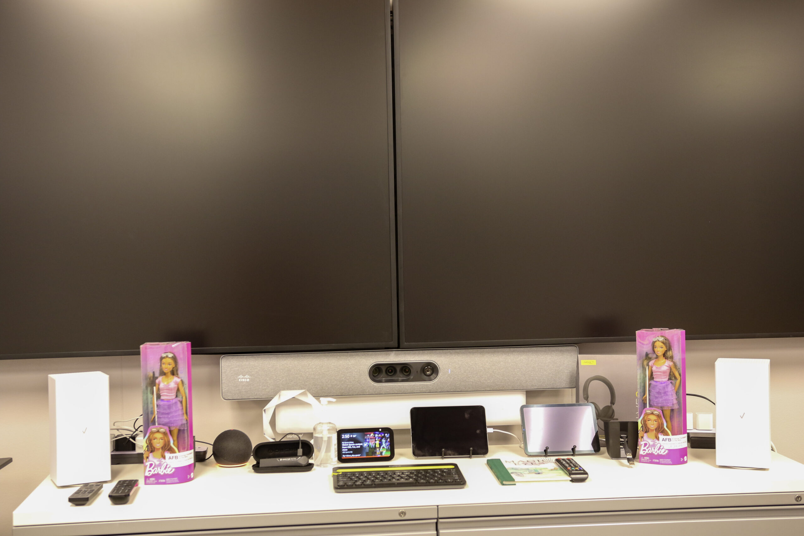 Table with Fios Routers, OTT devices: Fire TV Cube, Amazon Alexa devices, Mobile Device platform: iOS & Android, and two Barbie dolls with white canes