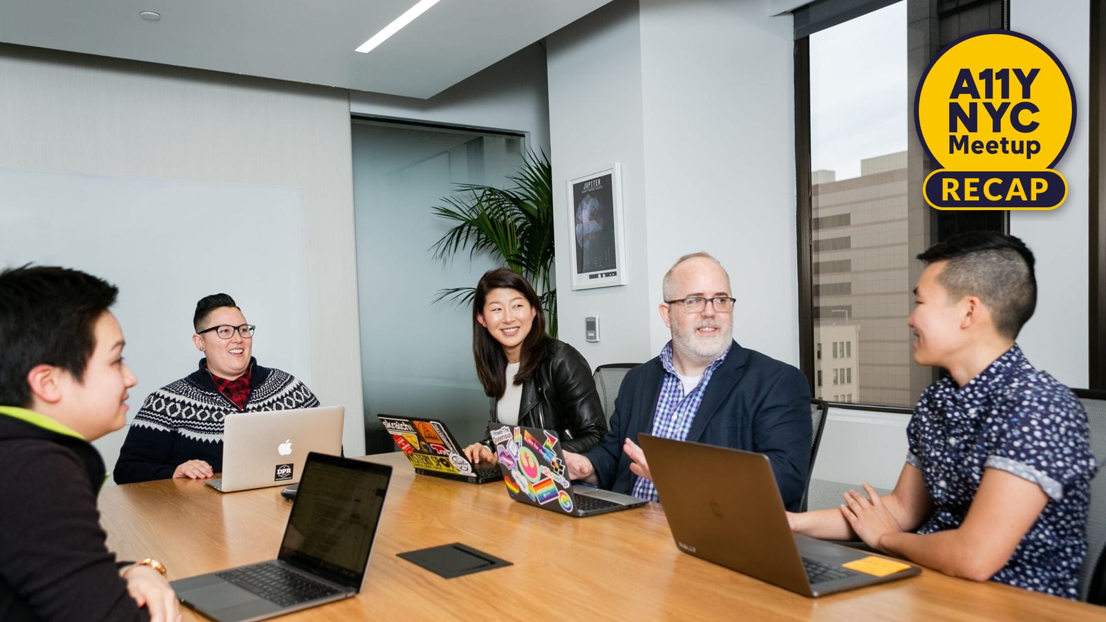 Five people gathered around a conference table in a modern office, engaged in conversation and using laptops. The group is diverse in age, gender, and style, with expressions suggesting engagement and collaboration.