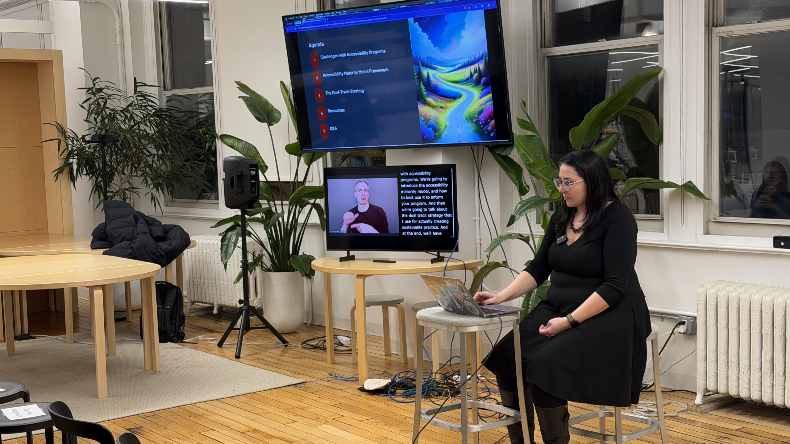 Lina sits on a stool in front of a laptop with a big screen to the side showing the presentation. Below is a smaller screen showing the ASL interpreter and captions.