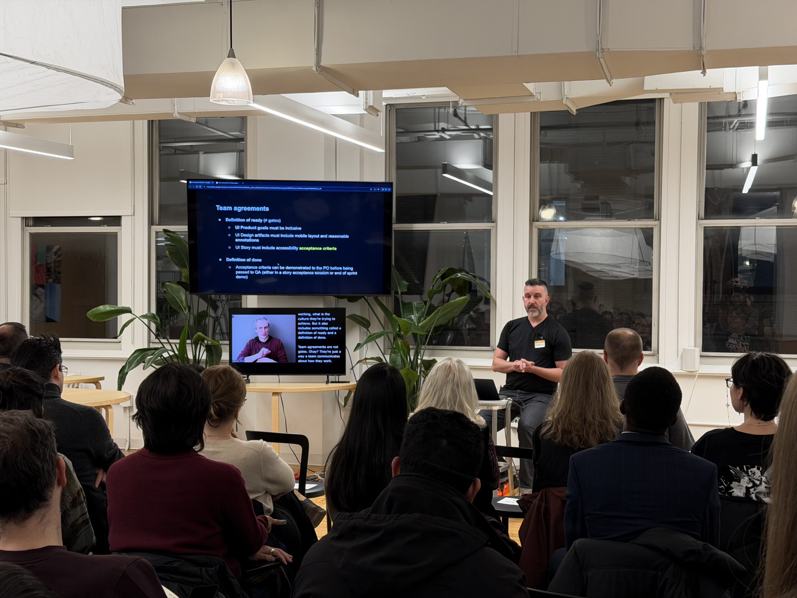 Charlie Triplett sits on a stool in front of a laptop with a big screen to the side showing the presentation. Below is a smaller screen showing the ASL interpreter and captions.