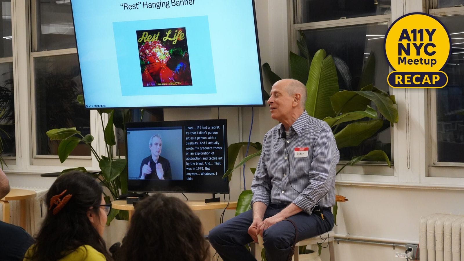 Peter Slatin sits on a stool next to a large screen with his slides and below the screen is a smaller screen displaying an ASL interpreter and captions.