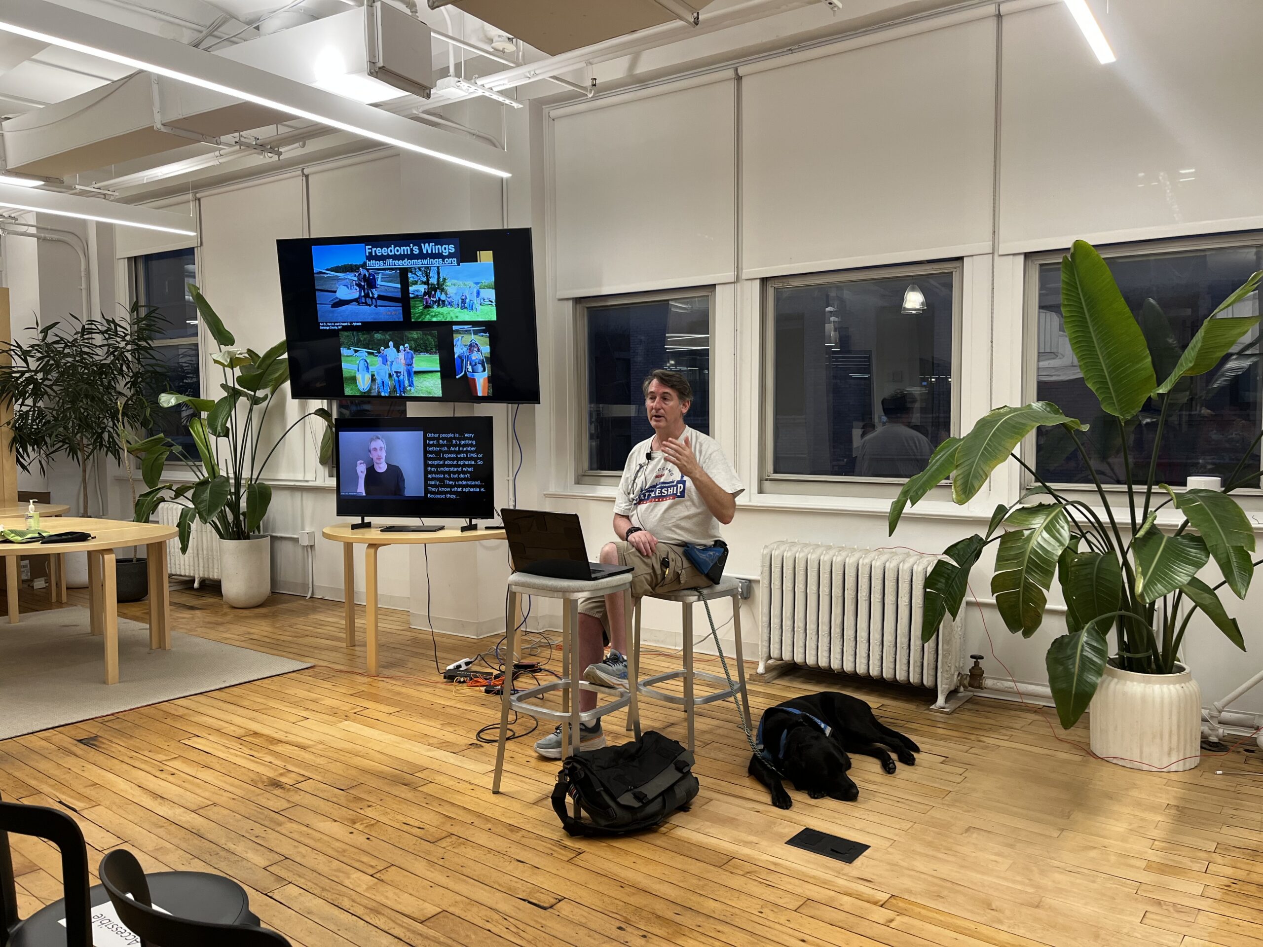 Avi Golden sits on a stool presenting. A service dog lies on the floor next to him. Two monitors next to Avi; one shows his slides and the other contains captions and an ASL interpreter.